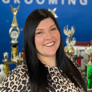 Katelyn Smith, CMG, professional dog grooming instructor and mentor with paragon pet school, shown in front of Best In Show trophies she's won